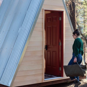 Puede incluir: Una persona camina hacia una peque&ntilde;a caba&ntilde;a de madera en forma de A con una puerta roja. La caba&ntilde;a tiene un techo de metal y est&aacute; rodeada de &aacute;rboles.
