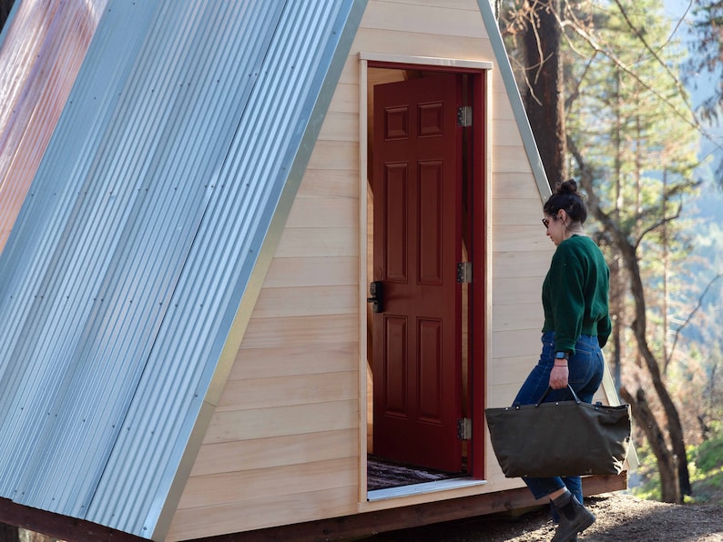 Puede incluir: Una persona camina hacia una peque&ntilde;a caba&ntilde;a de madera en forma de A con una puerta roja. La caba&ntilde;a tiene un techo de metal y est&aacute; rodeada de &aacute;rboles.