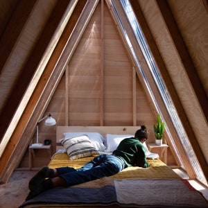 Puede incluir: Una persona yace en una cama en una caba&ntilde;a de madera con forma de A con una gran ventana. La cama tiene una colcha amarilla y marr&oacute;n y una manta verde. La persona lleva pantalones vaqueros azules y un jersey verde.