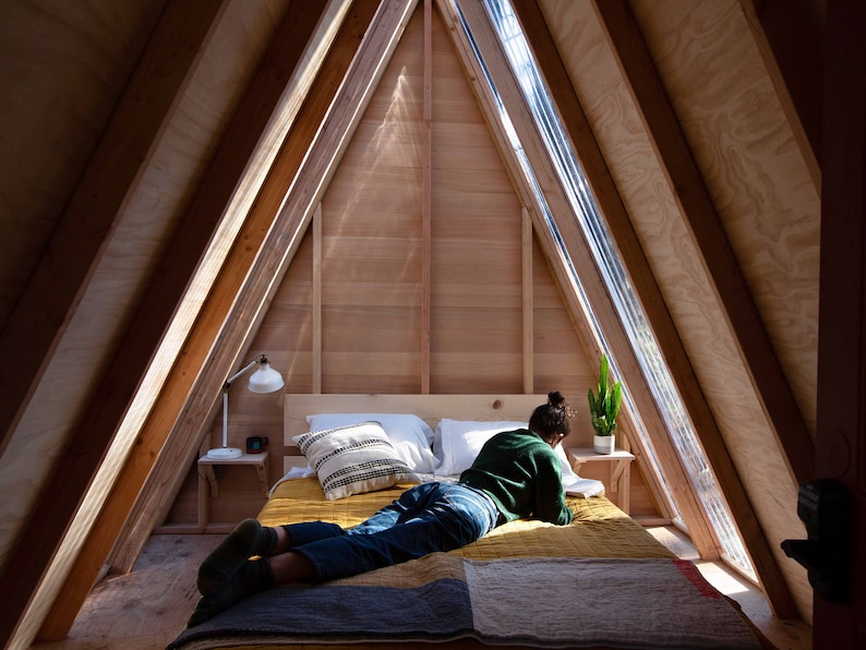 Pu&ograve; includere: Una persona giace su un letto in una cabina di legno a forma di A con una grande finestra. Il letto ha una trapunta gialla e marrone e una coperta verde. La persona indossa jeans blu e un maglione verde.