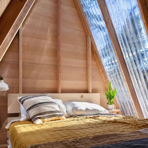 Puede incluir: Un dormitorio con una cama de estructura de madera con una colcha amarilla y almohadas blancas. La habitaci&oacute;n tiene un techo inclinado con grandes ventanas que dejan entrar la luz natural. Una peque&ntilde;a planta se encuentra en un estante junto a la cama.