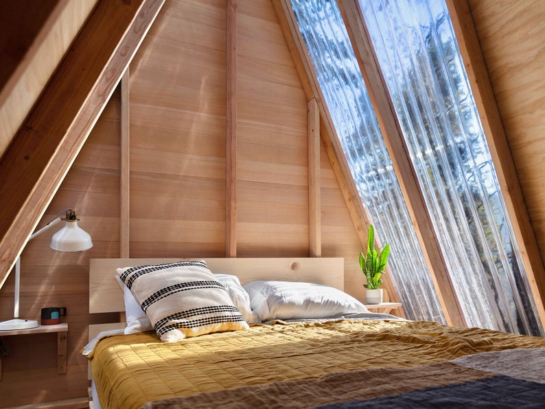 Puede incluir: Un dormitorio con una cama de estructura de madera con una colcha amarilla y almohadas blancas. La habitaci&oacute;n tiene un techo inclinado con grandes ventanas que dejan entrar la luz natural. Una peque&ntilde;a planta se encuentra en un estante junto a la cama.