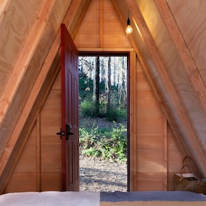 Puede incluir: Una caba&ntilde;a de madera en forma de A con una puerta roja abierta que revela una vista de un bosque. La caba&ntilde;a tiene un techo y paredes de madera, y una cama es visible en primer plano.