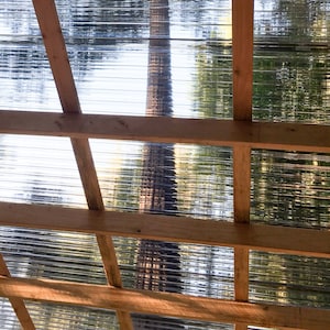 May include: A wooden frame with clear corrugated plastic panels, creating a roof structure. The panels allow light to filter through, creating a dappled effect on the ground below.