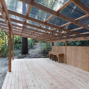 May include: A wooden deck with a wooden roof and a wooden fence. The roof is made of corrugated plastic panels. There is a wooden table and two stools on the deck.