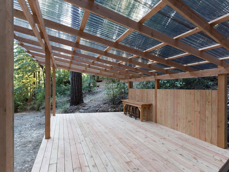 May include: A wooden deck with a wooden roof and a wooden fence. The roof is made of corrugated plastic panels. There is a wooden table and two stools on the deck.