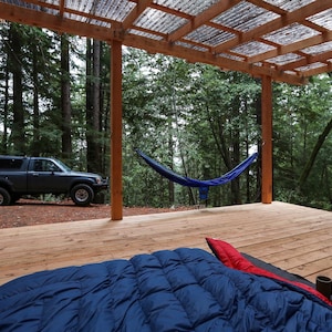 May include: A wooden deck with a blue hammock hanging between two wooden posts. A blue sleeping bag is on the deck with a mug on top of it. A gray pickup truck is parked in the background.