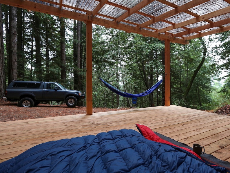 May include: A wooden deck with a blue hammock hanging between two wooden posts. A blue sleeping bag is on the deck with a mug on top of it. A gray pickup truck is parked in the background.