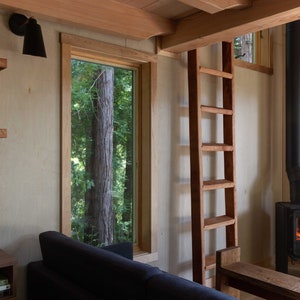 May include: A wooden ladder leading to a loft space in a cabin with a wood-burning stove and a window overlooking a forest.