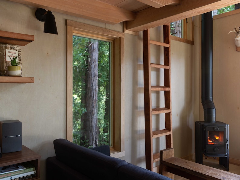 May include: A wooden ladder leading to a loft space in a cabin with a wood-burning stove and a window overlooking a forest.