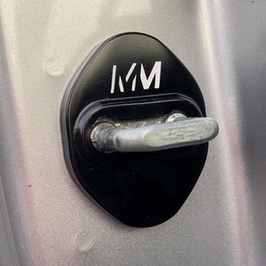 May include: Black car door latch cover with the white letters "MM". The cover is diamond-shaped and sits over the door latch. The background is a silver car door.