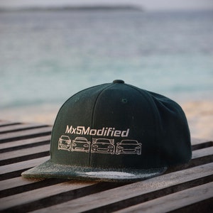 Puede incluir: Una gorra de béisbol verde oscuro con el texto "Mx5Modified" bordado en la parte delantera. La gorra presenta un contorno blanco de cinco modelos de coches diferentes.