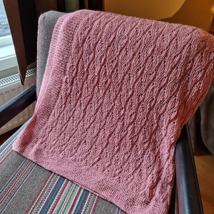 Peut inclure: Une couverture tricotée rose poussiéreux avec un motif de vagues détaillé est drapée sur une chaise rayée. La texture de la couverture est visible, mettant en valeur le travail de couture complexe. La couverture est d'une couleur douce et chaude.