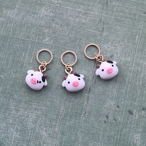 以下が含まれることがあります： 白い牛のチャームが付いたゴールドのステッチマーカーが3つ。各チャームには黒い斑点とピンクの鼻があります。