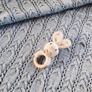 Peut inclure: Une couverture de bébé en tricot bleu clair avec un motif en tricot torsadé texturé. Un hochet en forme de lapin crocheté avec un anneau en bois est posé sur la couverture. L'ensemble est doux et accueillant.