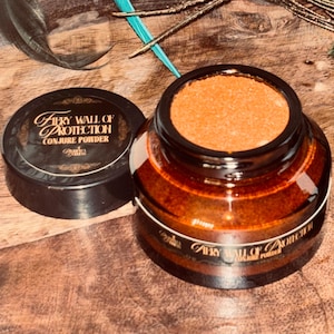 Op de afbeelding: Een kleine amberkleurige glazen pot gevuld met oranje poeder, vergezeld van een zwarte deksel. Het etiket op de pot en de deksel bevat de gouden tekst Fiery Wall of Protection Conjure Powder. De pot staat naast verschillende pauwenveren.