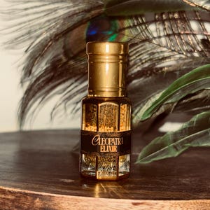 Może przedstawiać: Mała, ozdobna butelka perfum z złotą nakrętką i czarną etykietą z napisem "Cleopatra Elixir". Butelka jest wypełniona ciemnym płynem i stoi na drewnianej powierzchni, z piórami pawia w tle.