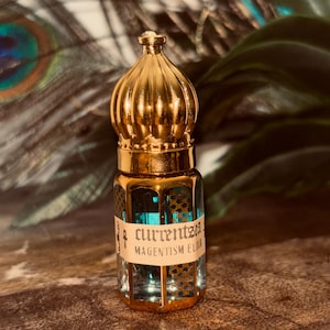May include: A small, ornate glass bottle with a gold-colored lid and accents. The bottle contains a teal-colored liquid and has a label that reads "currentsea MAGENTISM ELIXIR". The background features peacock feathers and greenery.