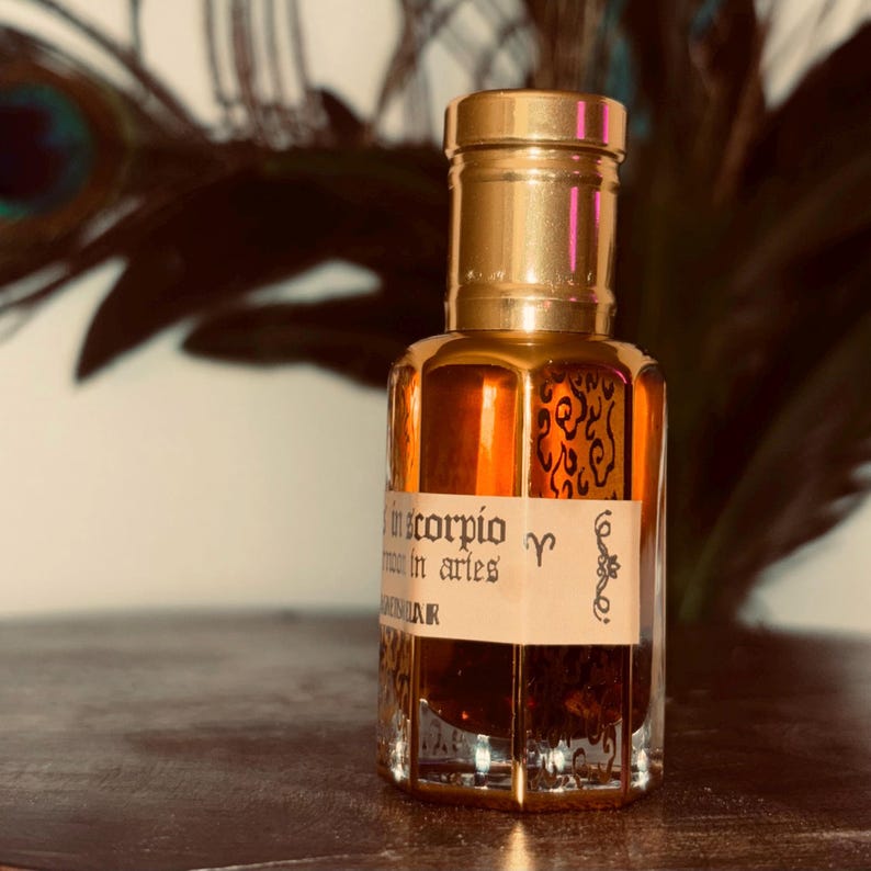 May include: A small, clear glass perfume bottle with a gold-colored cap and accents. The bottle contains a dark amber liquid and has a label with text and zodiac symbols. The background includes a blurred peacock feather.