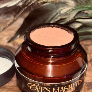 Op de afbeelding: Een bruine glazen pot met een donkerbruin deksel en een wit etiket met gouden tekst met de tekst "Eve's Magnetism Powder". De pot bevat een lichtbruin poeder. Het deksel is eraf en onthult een witte substantie. De achtergrond bevat groene en bruine veren.