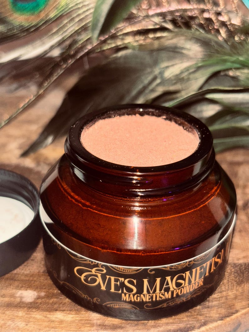Op de afbeelding: Een bruine glazen pot met een donkerbruin deksel en een wit etiket met gouden tekst met de tekst "Eve's Magnetism Powder". De pot bevat een lichtbruin poeder. Het deksel is eraf en onthult een witte substantie. De achtergrond bevat groene en bruine veren.