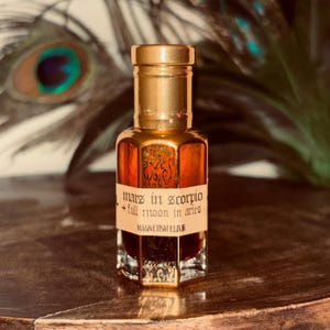 May include: A small, ornate, amber-colored glass bottle with a gold cap. The bottle contains a dark liquid and has a label that reads "Mars in Scorpio + Full Moon in Aries MAGNETISM ELIXIR". The background features a peacock feather.