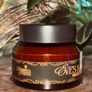 Op de afbeelding: Een potje van het product Eve's Mask Magnetism met een zwart deksel en een donkerbruine glazen pot. Het etiket is zwart met gouden letters en een decoratieve rand. De merknaam Divine Lea Goddess staat ook op het etiket.