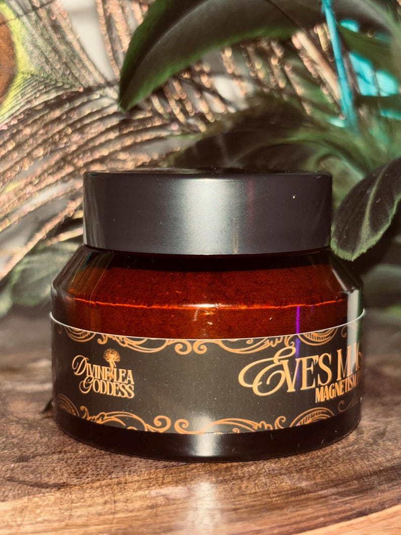 Op de afbeelding: Een potje van het product Eve's Mask Magnetism met een zwart deksel en een donkerbruine glazen pot. Het etiket is zwart met gouden letters en een decoratieve rand. De merknaam Divine Lea Goddess staat ook op het etiket.