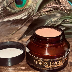 Op de afbeelding: Een bruine glazen pot met een zwarte deksel, met een lichtbruin poeder. De pot heeft een etiket met de tekst "Eve's Magnetism Powder" in gouden letters. De achtergrond bevat pauwenveren.