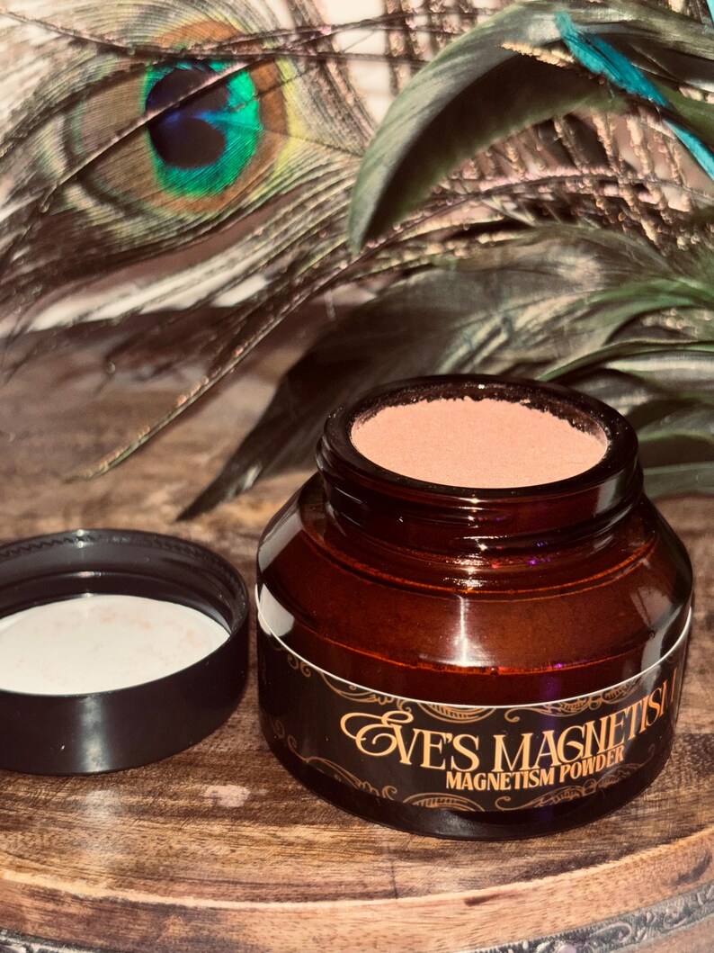 Op de afbeelding: Een bruine glazen pot met een zwarte deksel, met een lichtbruin poeder. De pot heeft een etiket met de tekst "Eve's Magnetism Powder" in gouden letters. De achtergrond bevat pauwenveren.