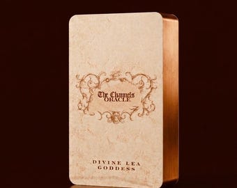 May include: A rectangular oracle card deck with rounded corners. The front of the deck features the text "The Channels Oracle" in a decorative frame, with "Divine Lea Goddess" below. The deck has a textured, off-white front and copper-colored edges.