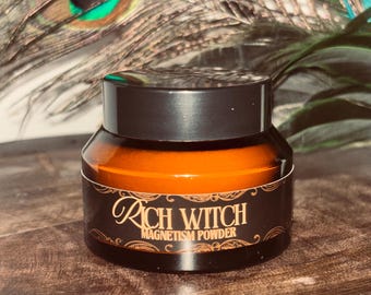 May include: A small amber glass jar with a black screw-top lid. The label features decorative gold-toned borders and the text "RICH WITCH MAGNETISM POWDER" in a stylized font. The jar is cylindrical with a wide base and a tapered neck.