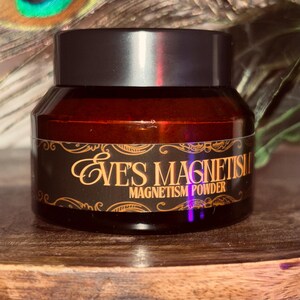Op de afbeelding: Een kleine, donkere glazen pot met een zwart deksel. De pot heeft een etiket met de woorden "EVE'S MAGNETISM MAGNETISM POWDER" in gouden letters. De pot staat op een houten oppervlak.