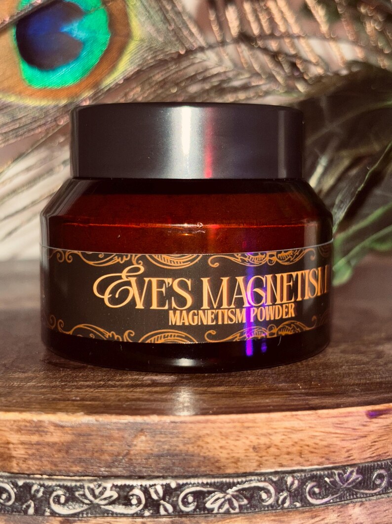Op de afbeelding: Een kleine, donkere glazen pot met een zwart deksel. De pot heeft een etiket met de woorden "EVE'S MAGNETISM MAGNETISM POWDER" in gouden letters. De pot staat op een houten oppervlak.
