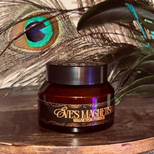 Op de afbeelding: Een kleine, bruine glazen pot met een zwart deksel, met het opschrift "Eve's Magnetism Magnetism Powder" in gouden letters. De pot staat op een houten oppervlak, met pauwenveren op de achtergrond.