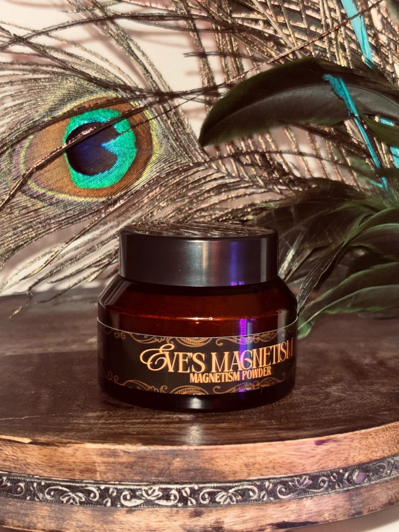 Op de afbeelding: Een kleine, bruine glazen pot met een zwart deksel, met het opschrift "Eve's Magnetism Magnetism Powder" in gouden letters. De pot staat op een houten oppervlak, met pauwenveren op de achtergrond.