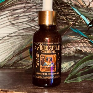 May include: A dark brown glass bottle of "Vindicate Me Conjure Oil" with a gold cap and white dropper. The label features ornate script and an illustration of a person. Text includes "Justice, Karma, Law Go Away" and "Conjured with Jupiter and Mars."