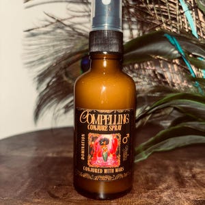 Pode incluir: Uma garrafa de vidro castanho de "Compelling Conjure Spray" com uma tampa preta e um pulverizador. O rótulo apresenta as palavras "Conjured with Mars" e um gráfico. A garrafa está sobre uma superfície de madeira, com penas de pavão no fundo.