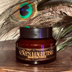 Op de afbeelding: Een kleine, donkerbruine pot met een zwart deksel, met het opschrift "EVE'S MAGNETISM MAGNETISM POWDER" in gouden letters. De pot staat op een houten oppervlak, met pauwenveren op de achtergrond.