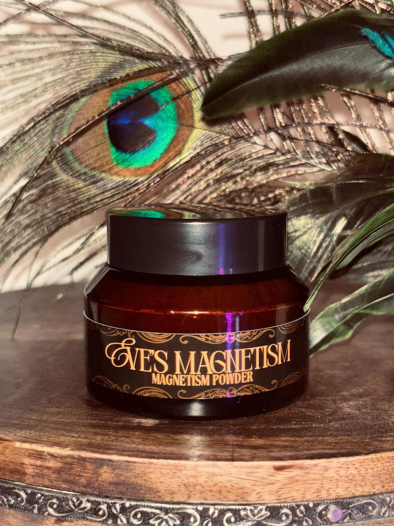 Op de afbeelding: Een kleine, donkerbruine pot met een zwart deksel, met het opschrift "EVE'S MAGNETISM MAGNETISM POWDER" in gouden letters. De pot staat op een houten oppervlak, met pauwenveren op de achtergrond.