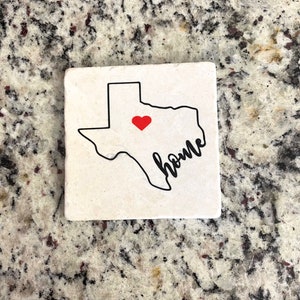 Può includere: Un sottobicchiere bianco con un contorno nero dello stato del Texas. All'interno del contorno c'è un cuore rosso e la parola "home" scritta in corsivo nero.
