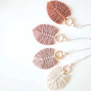May include: Four handmade macrame leaf ornaments with wooden rings and string. The leaves are in shades of brown, pink, and white.