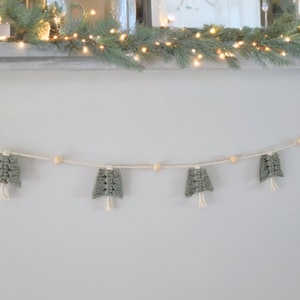 May include: A handmade Christmas tree garland. The garland features small, grey macrame Christmas trees with white fringe, strung along a white cord. Wooden beads are interspersed between the trees. The garland is displayed against a white wall.
