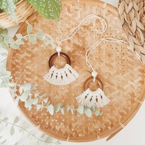 May include: Two wooden ring teethers with white tassel fringe hanging from a woven bamboo tray. The tray is surrounded by green eucalyptus branches.
