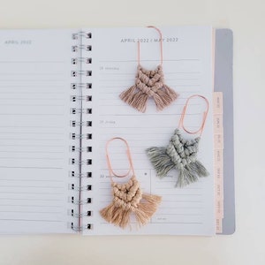 May include: Three macrame bookmarks with fringe, each attached to a rose gold paper clip. The bookmarks are in shades of beige and green. The bookmarks are on a white page with the text "APRIL 2022 / MAY 2022".