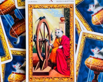 Può includere: Carte dei tarocchi a tema gatto. La carta centrale raffigura un gatto in abito rosso che aziona una ruota, circondato da altre carte con un gatto in un calice d'oro. Le carte hanno bordi dorati su sfondo blu.