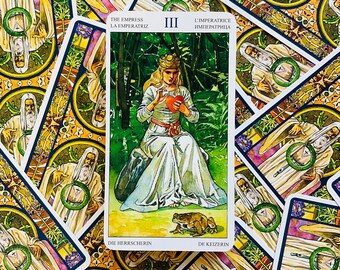 Può includere: La carta dei tarocchi L'Imperatrice del mazzo Rider-Waite. La carta mostra una donna con un vestito bianco seduta su una roccia in un bosco. Tiene un cuore rosso in mano. Una rana è seduta a terra di fronte a lei. La carta è circondata da altre carte dei tarocchi.