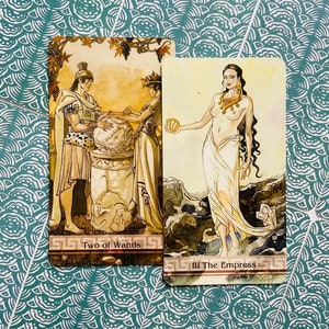 May include: Two tarot cards, "Two of Wands" and "III The Empress". The Two of Wands card depicts two figures standing on a platform, one holding a globe and the other holding a staff. The Empress card shows a woman standing on a rocky shore, holding a fruit in her hand. Both cards have a gold background with intricate details.