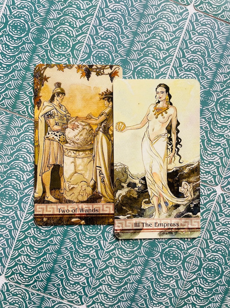 May include: Two tarot cards, "Two of Wands" and "III The Empress". The Two of Wands card depicts two figures standing on a platform, one holding a globe and the other holding a staff. The Empress card shows a woman standing on a rocky shore, holding a fruit in her hand. Both cards have a gold background with intricate details.