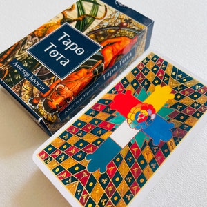 Pu&ograve; includere: Una scatola di mazzo di tarocchi con uno sfondo blu e un'illustrazione colorata di un toro e un uomo a cavallo. Il testo "Taro Tota" &egrave; stampato in bianco su uno stendardo blu. Il testo "Alister Crowley" &egrave; stampato in bianco su uno stendardo blu. La scatola &egrave; aperta ed &egrave; visibile una carta dei tarocchi. La carta ha uno sfondo bianco con un design colorato di una croce a quattro braccia, ogni braccio di un colore diverso. La croce &egrave; circondata da uno schema di diamanti neri e dorati.